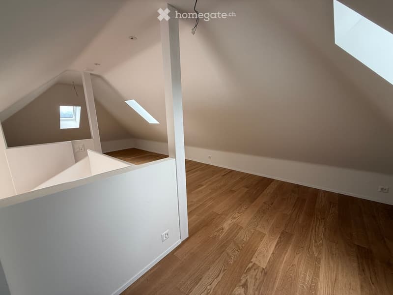 An empty attic space with wooden flooring, white walls, and skylights providing natural light. The room features exposed wooden beams and a small raised platform area.