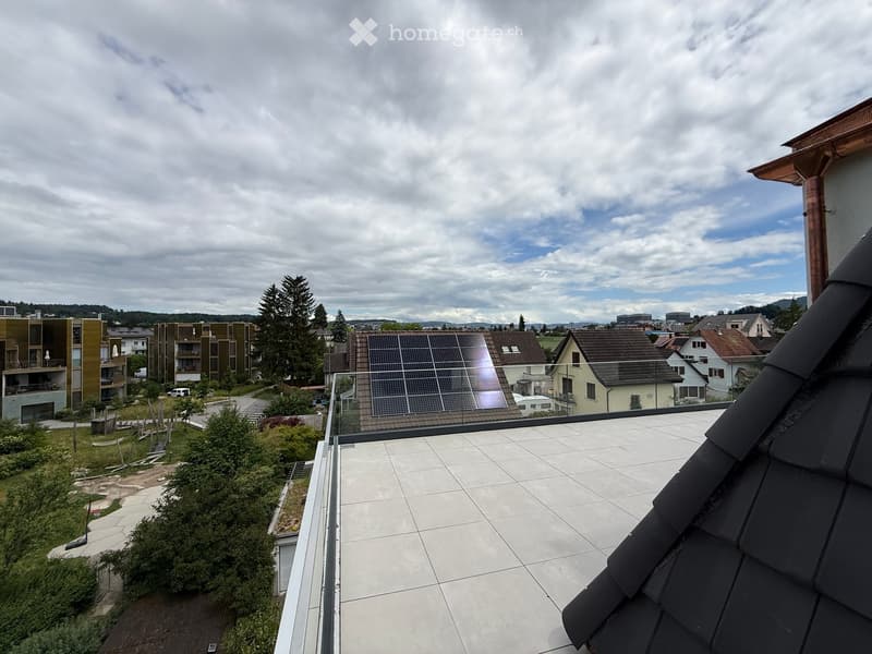 Roof terrace with tiled flooring, glass railing, and a view of a residential area with houses, trees, and solar panels on one roof.