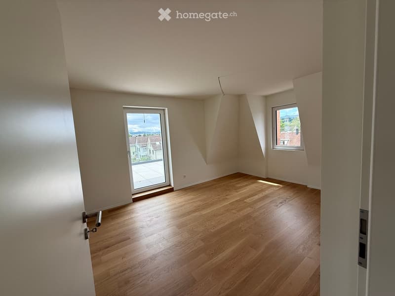 Empty room with wooden flooring, white walls, and large windows offering a view of the outside. The room has a sliding glass door leading to a balcony.