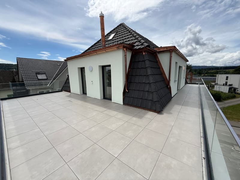 Roof terrace with light-colored tiles, glass railing, and access to a modern house with a distinctive roofline and chimney.