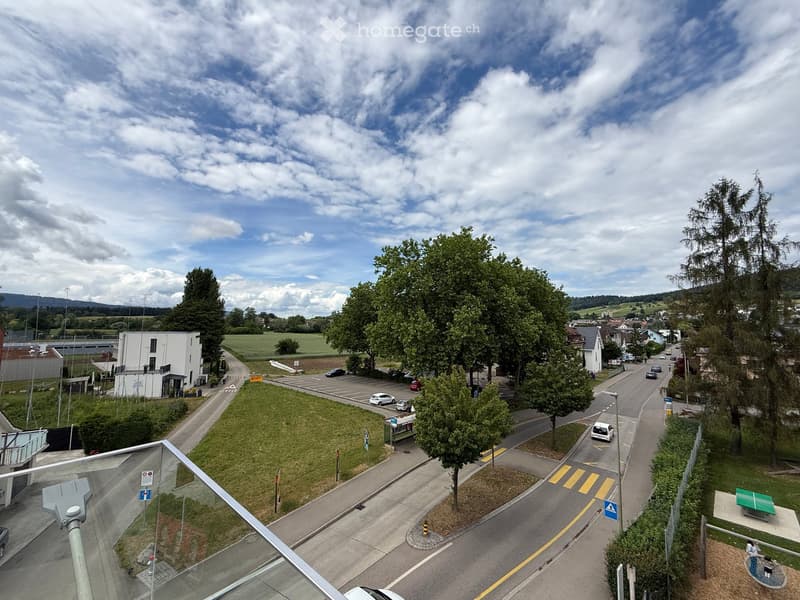 A view from a balcony or terrace overlooking a suburban area with roads, parked cars, trees, and a small park area with a green table and benches.