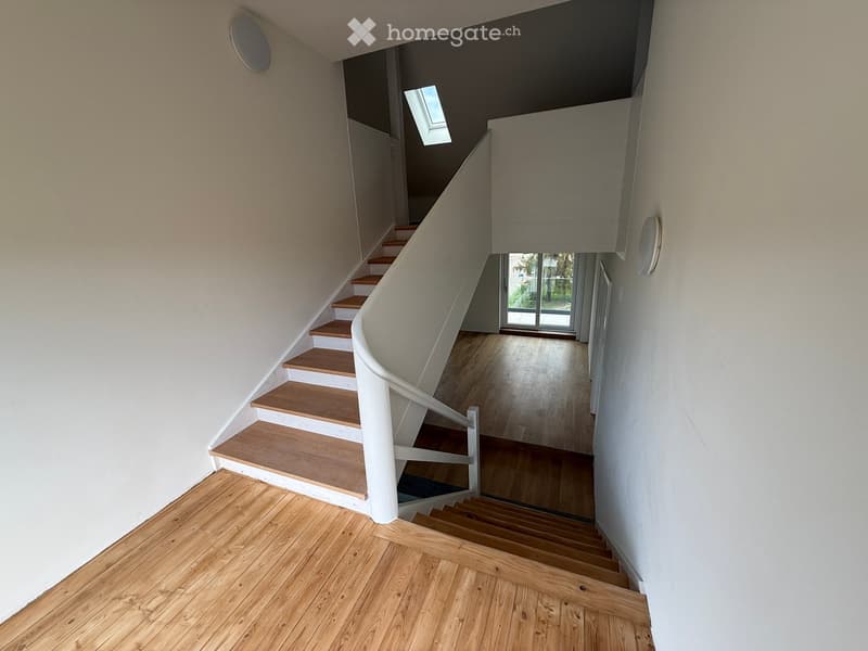 Spacious hallway with wooden flooring, curved staircase with wooden steps and white railing, skylight window, and a view to an adjacent room with large glass doors.