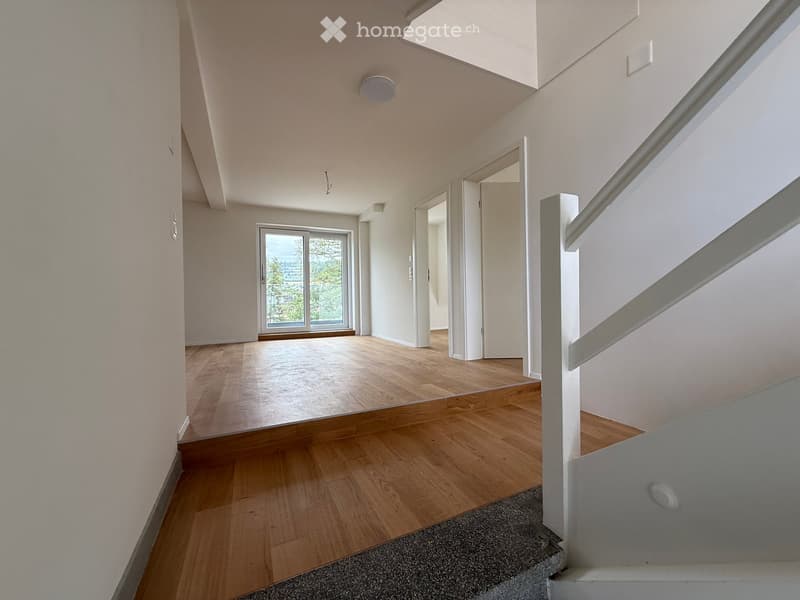 Bright, modern hallway with wooden flooring, white walls, and a staircase leading upwards. Features a large window at the end, allowing natural light to fill the space.