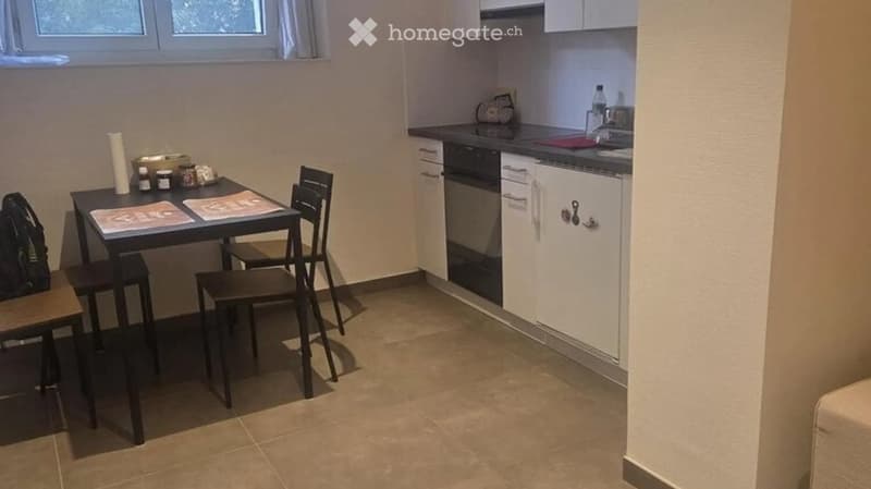 A compact kitchen with a small dining area. The kitchen features white cabinets, a black oven, and a dishwasher. A small black table with two chairs is placed near the window, and various items such as a backpack, jars, and a bottle are visible on the cou