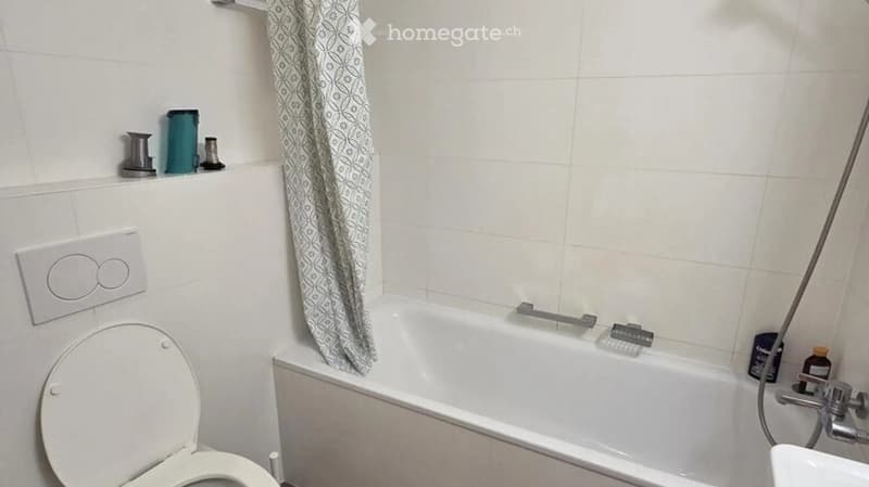 Bathroom with a toilet, bathtub, and shower curtain. The bathtub has a handheld shower attachment, and there are various toiletries on the shelf above the bathtub.