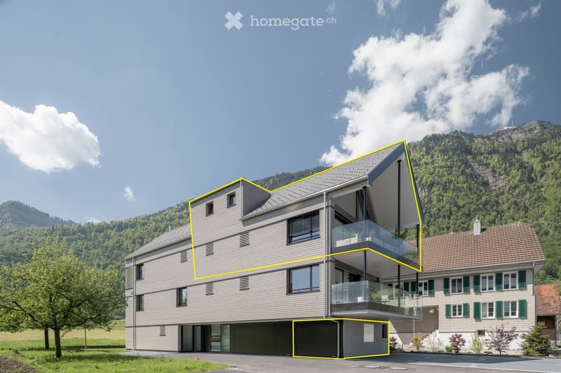Modern two-story house with grey siding, large windows, and a balcony on the upper level, set against a scenic mountain backdrop.