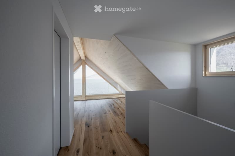 Bright, minimalist hallway with wooden flooring, white walls, and a skylight ceiling allowing natural light to flood the space. A window on the right side offers a view of the outdoors.