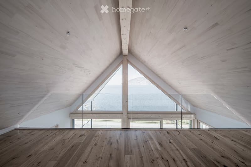 An empty room with wooden flooring and a wooden slanted ceiling, featuring a large glass railing and windows that offer a scenic view.