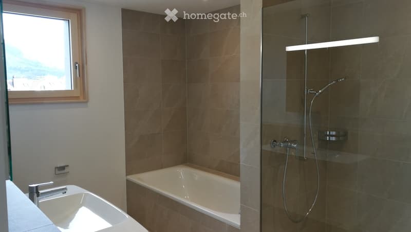 Modern bathroom with a bathtub, sink, and glass-enclosed shower. Features a large window with a scenic view, sleek fixtures, and warm brown tiles.