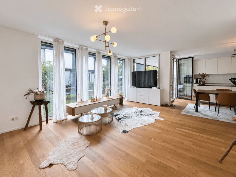 Modern living and dining area with hardwood flooring, large windows, a white sofa, glass coffee tables, a dining table with chairs, a television, and a kitchen area with cabinets.