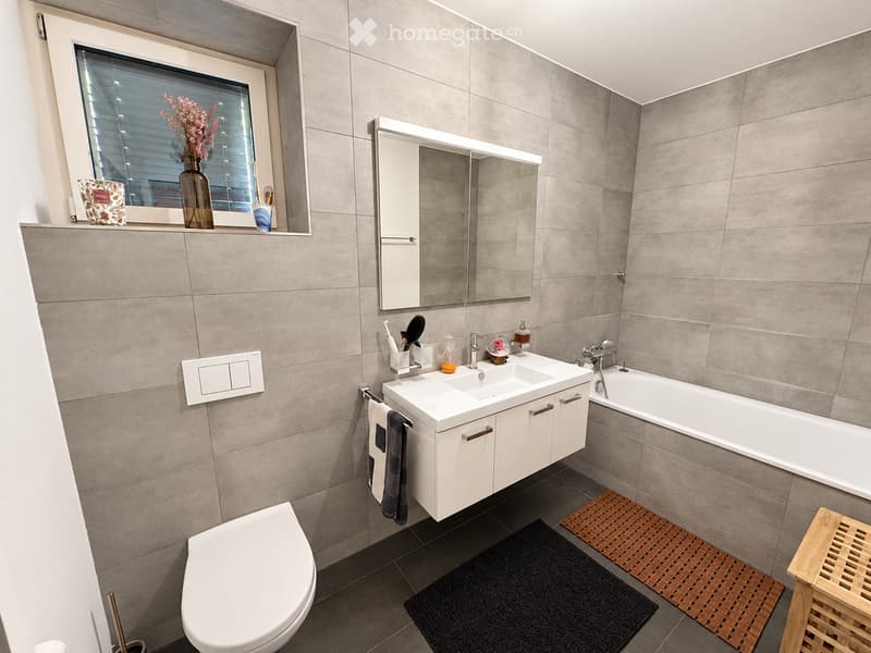 Modern bathroom with grey tiled walls, a double sink vanity with a mirror, a toilet, a bathtub, and a window with blinds. Includes towels, a vase with flowers, and a small wooden stool.
