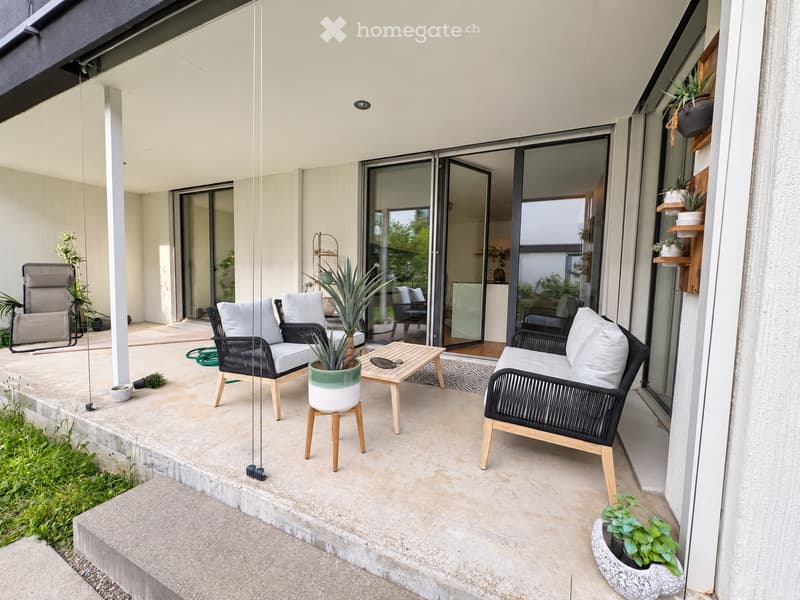 Modern balcony with a concrete floor, large sliding glass doors, and outdoor furniture including chairs, a small table, and potted plants.