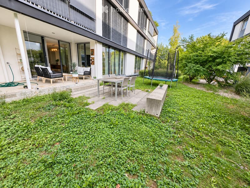Garden area with a modern house in the background, featuring a patio with outdoor furniture, a trampoline, and lush greenery.