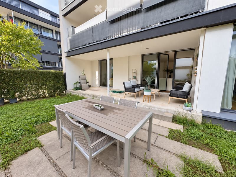 Modern outdoor patio with a wooden dining table, wicker chairs, and lounge chairs, surrounded by greenery and a modern building.
