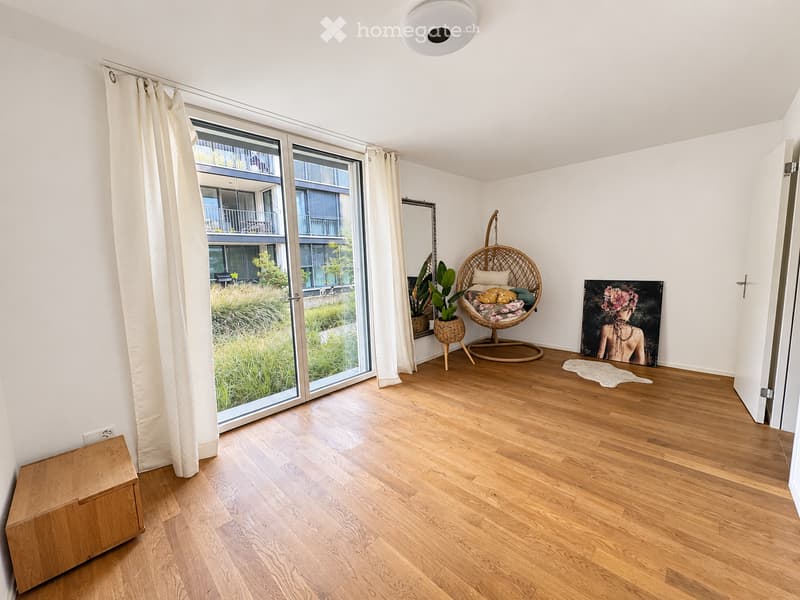 Bright living space with wooden flooring, large windows offering a view, a wicker hanging chair, potted plants, artwork, and a wooden storage box.