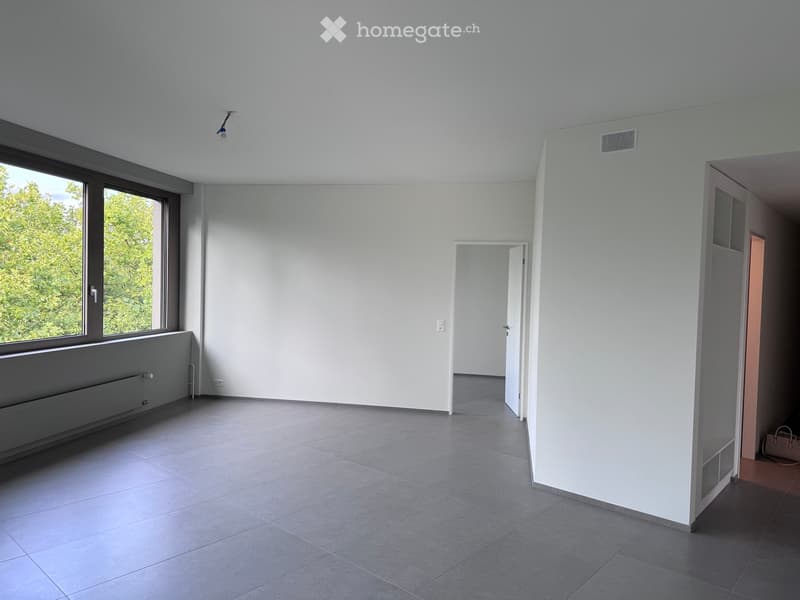 Empty room with white walls, grey tiled floor, large window with view of greenery, radiator, and open doorway leading to another space.