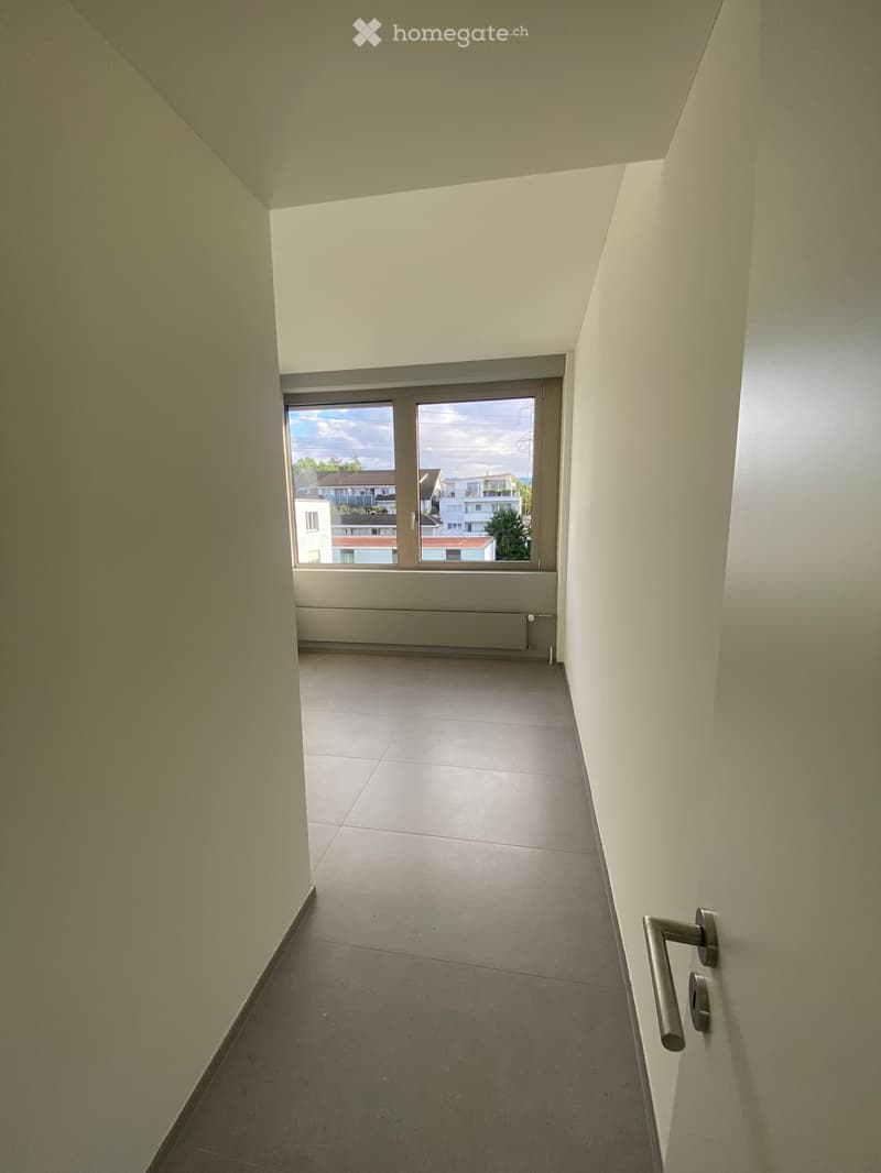 Empty hallway with light-colored walls, tiled floor, and a large window at the end showing a view of buildings and trees outside.