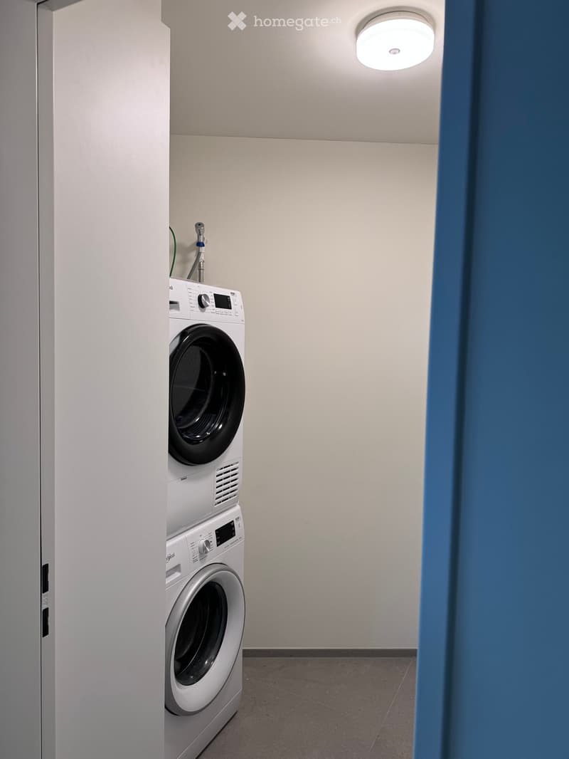 A small, well-lit room with a white washing machine and dryer stacked vertically against a wall.