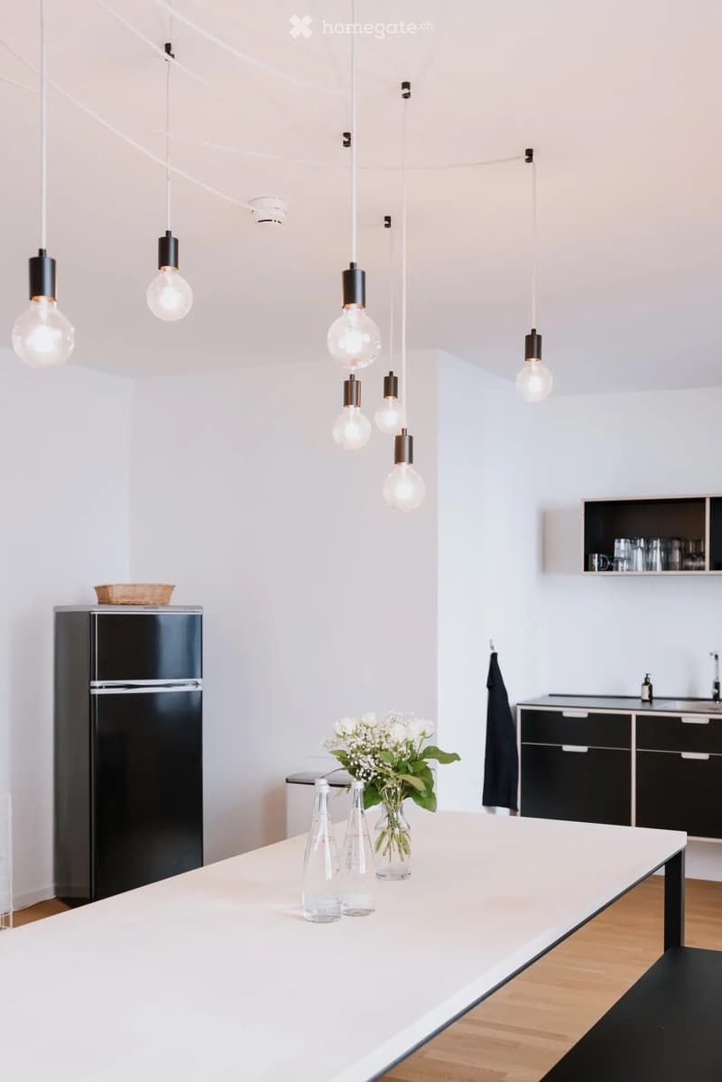 Modern kitchen with white countertops, black cabinets, and pendant lights. Features a refrigerator, a vase with white flowers, and several glass bottles on the counter.