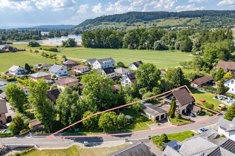 Aerial view of a residential neighborhood with multiple houses, surrounded by green fields, trees, and a river in the background.