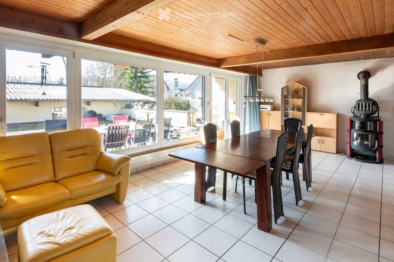 Bright living and dining area with a yellow leather sofa, ottoman, wooden dining table, chairs, and a wood-burning stove. Large windows provide a view of the outdoor patio.
