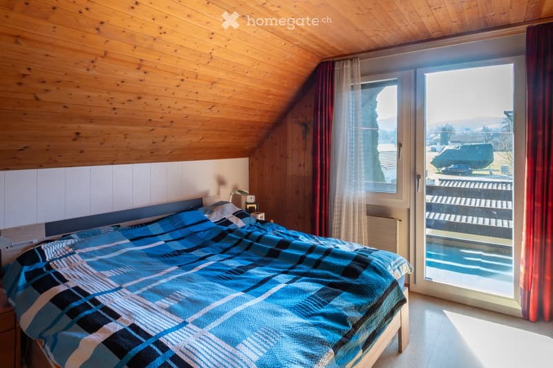 Cozy bedroom with wooden ceiling and walls, large window offering a scenic view, neatly made bed with blue and white plaid blanket, nightstand with lamp.