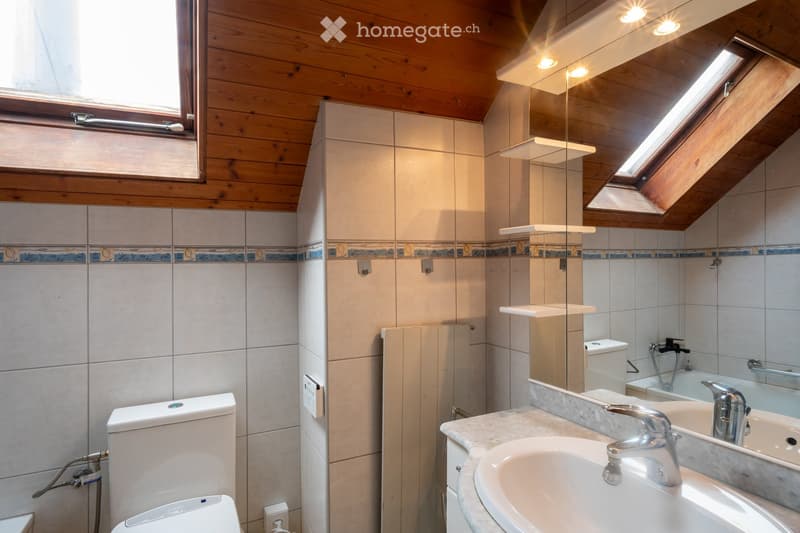 Modern bathroom with wooden ceiling, white tiles, and blue decorative border. Features a toilet, sink with chrome faucet, mirror, and bathtub.