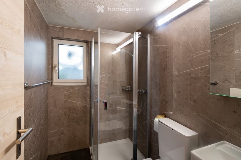 Modern bathroom with marble walls, a glass-enclosed shower, a toilet, a sink, and a mirror. Features include a frosted window and built-in lighting.