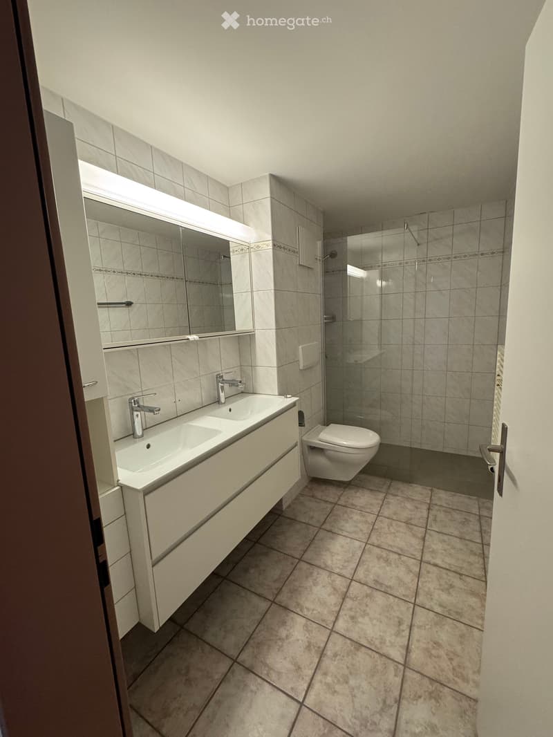 Modern bathroom with beige tiles, double sink, large mirror, and glass-enclosed shower area.