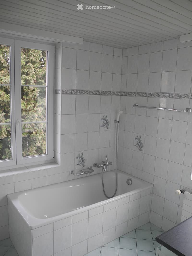 Bathroom with a white bathtub, tiled walls, a window with a view of greenery, and a showerhead attached to the wall.