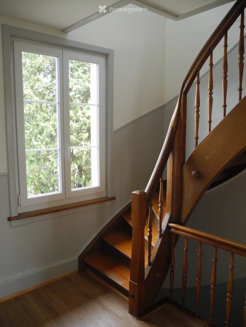 Wooden staircase with a handrail and balusters, leading up to a landing with windows showing greenery outside.