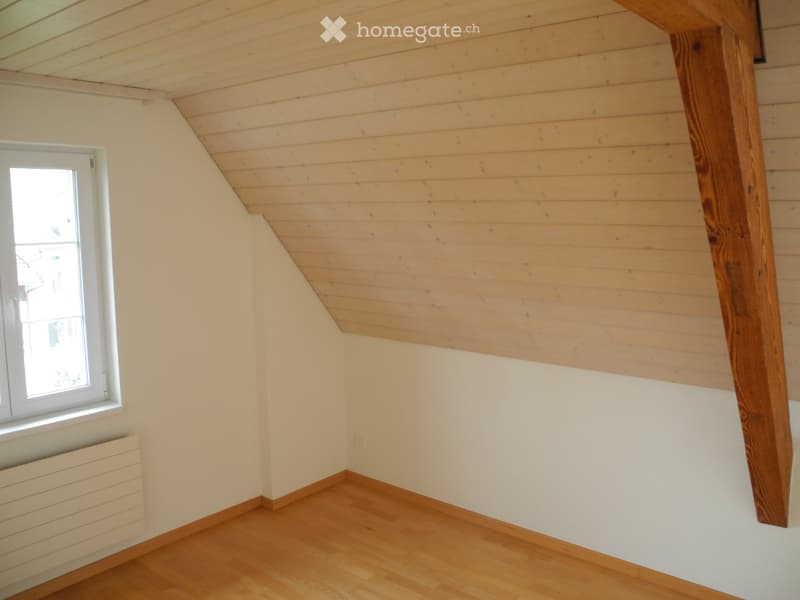 Empty attic room with wooden slat ceiling, wooden floor, white walls, and a single window with white frame.