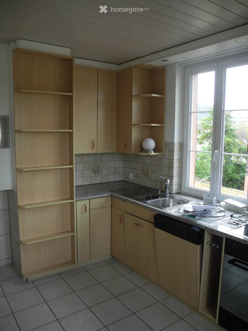 Modern kitchen with light wooden cabinets, granite countertop, stainless steel sink, and built-in dishwasher. The space features tiled walls and a window offering a view of greenery.