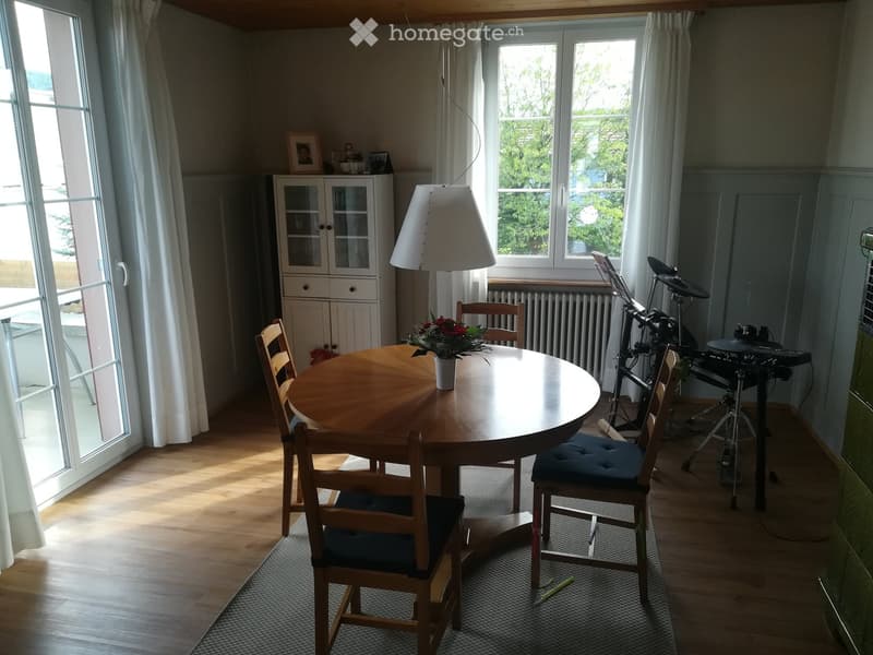 Brightly lit room with wooden flooring, large windows, a round wooden dining table with four chairs, a drum set, a white cabinet, and a hanging lamp.