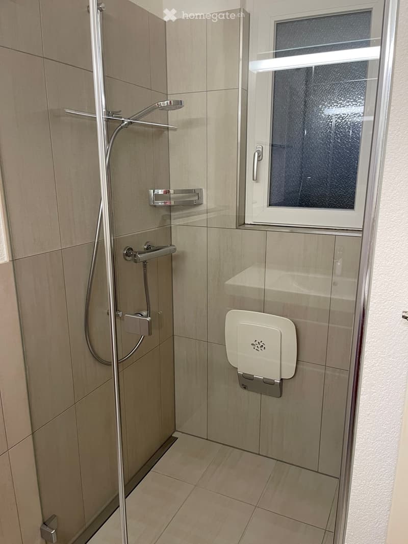 Modern shower area with beige tiled walls and floor, chrome showerhead and rail, towel rack, and toilet paper holder.