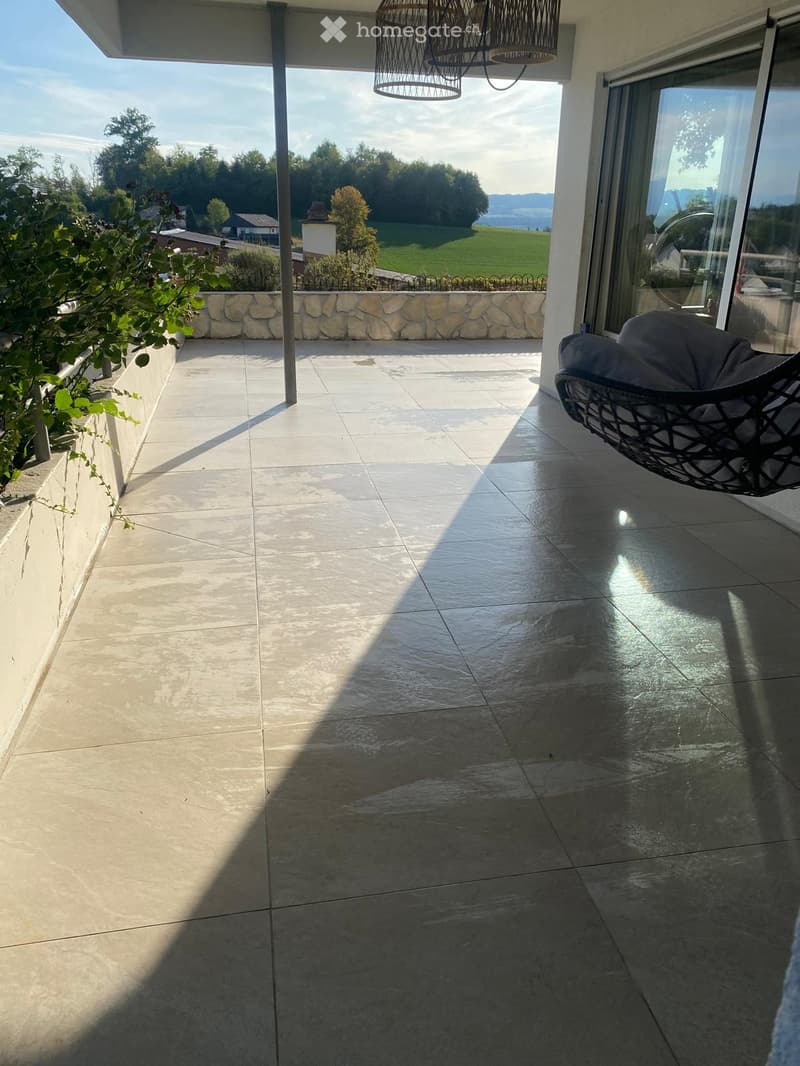 Balcony with tiled floor, wicker hanging chair, stone wall, and scenic countryside view.