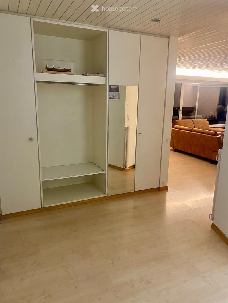 A furnished room with a wooden floor, a large closet with mirrored doors, and a visible living area with a sofa.
