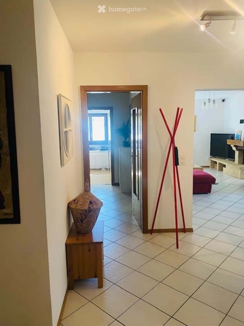 A narrow hallway with tiled flooring, white walls, and a wooden door leading to another room. A small wooden bench with a decorative vase is placed against the wall, and a red coat rack stands nearby.