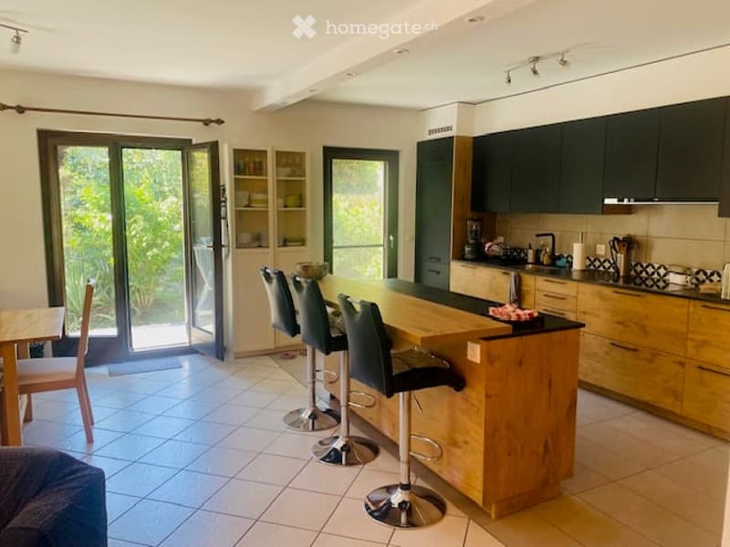 Modern kitchen with wooden island, four stools, dark green cabinets, and various appliances including a dishwasher and coffee maker. Large sliding glass doors lead to an outdoor area.