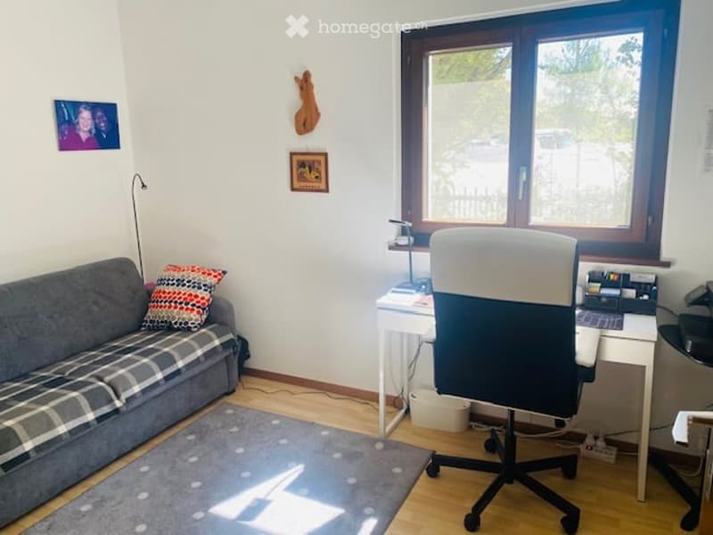 A cozy, well-lit room with a grey sofa, a patterned throw pillow, a white desk with a chair, a floor lamp, and a picture frame on the wall.