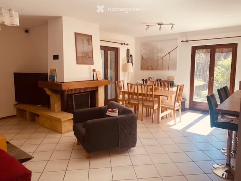A modern living room with a fireplace, a sofa, a dining table with chairs, and a lamp. The room features large windows and a tiled floor.