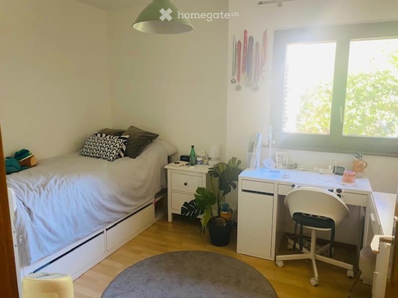 Cozy bedroom with a single bed, nightstand, desk, chair, and a small plant. The room features a round rug and a hanging green lamp.