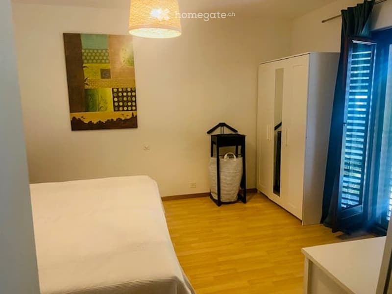 Cozy bedroom with a wooden floor, a bed, a wardrobe with a mirror, a hanging lamp, and a colorful painting on the wall.