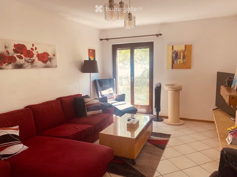 Bright living room with red sectional sofa, coffee table, armchair, floor lamp, and cat tree. Large sliding glass door offers a view of greenery outside.