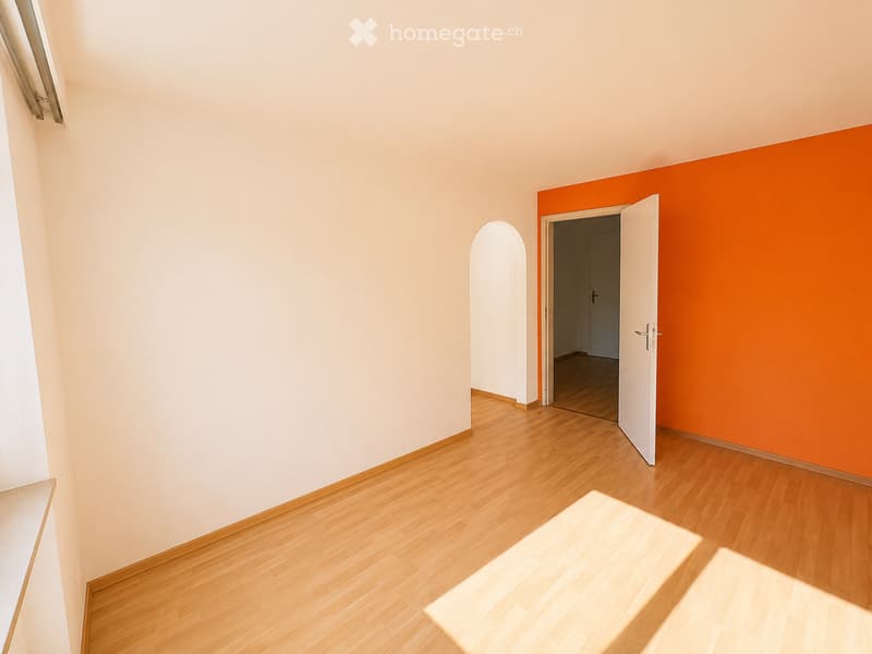 Empty room with light wooden flooring and white walls, featuring an open doorway leading to another room with orange accent wall.