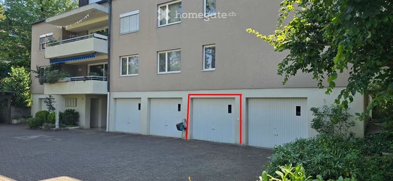 Multi-story residential building with balconies, multiple garage doors, and surrounding greenery.
