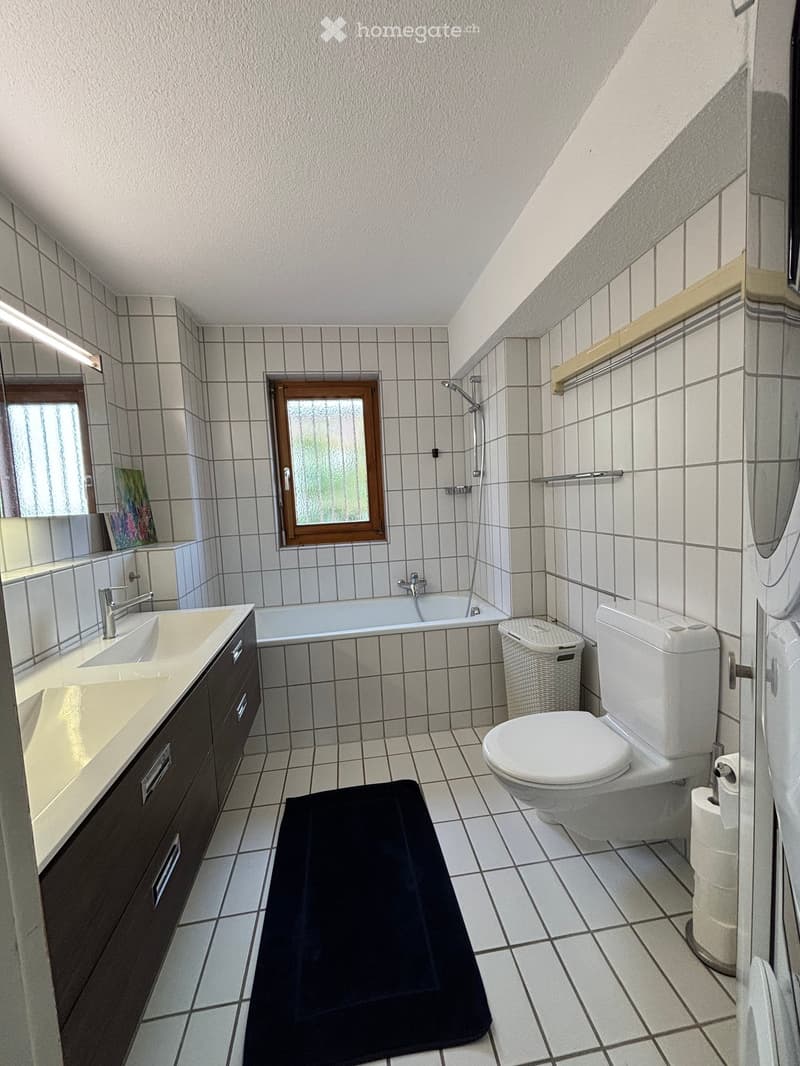Bathroom with white tiled walls and floor, double sink with dark wood cabinetry, bathtub, toilet, wicker basket, and a black bath mat.