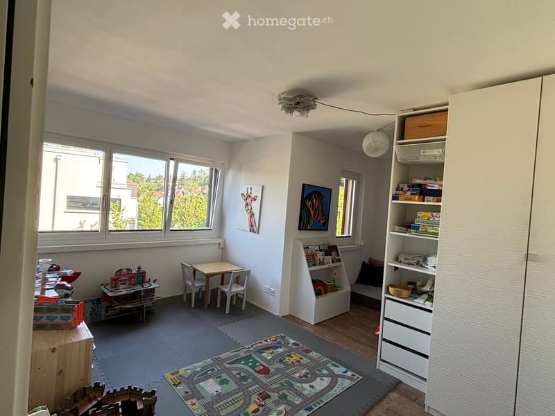 Brightly lit children's room with large windows, a small table and chairs, a bookshelf, and colorful toys.