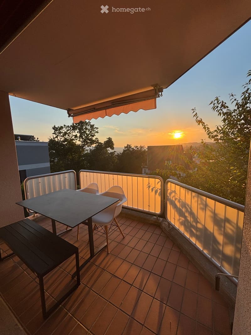 Balcony with tiled floor, metal railing, table, and chairs, overlooking a cityscape at sunset.