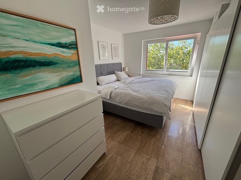 Modern bedroom with a large bed, nightstand, lamp, and artwork on the wall. The room features a large window with a view of greenery outside.